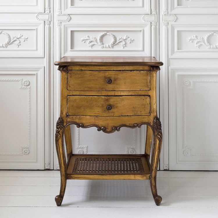 Vintage wooden side table with two drawers, ornate legs, and a lower woven shelf, set against an elegant white paneled wall.