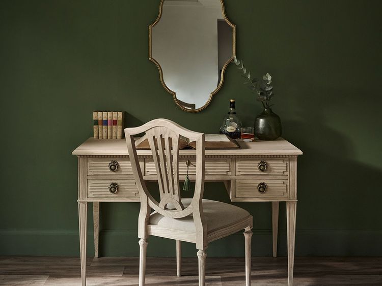 Elegant wooden desk with a chair, mirror, books, plant, and a decanter on a dark green wall background.