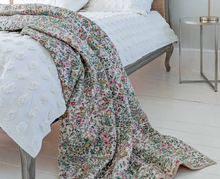 A cosy bedroom with a white textured bedspread, a colourful floral quilt draped over the side, and a round side table on a white wooden floor.