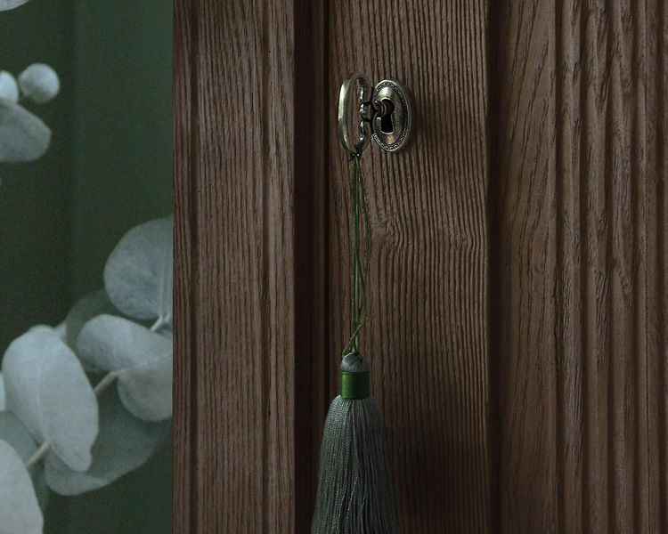 A wooden door with a vintage keyhole, adorned with a green tassel. A blurred leafy pattern is visible in the background.