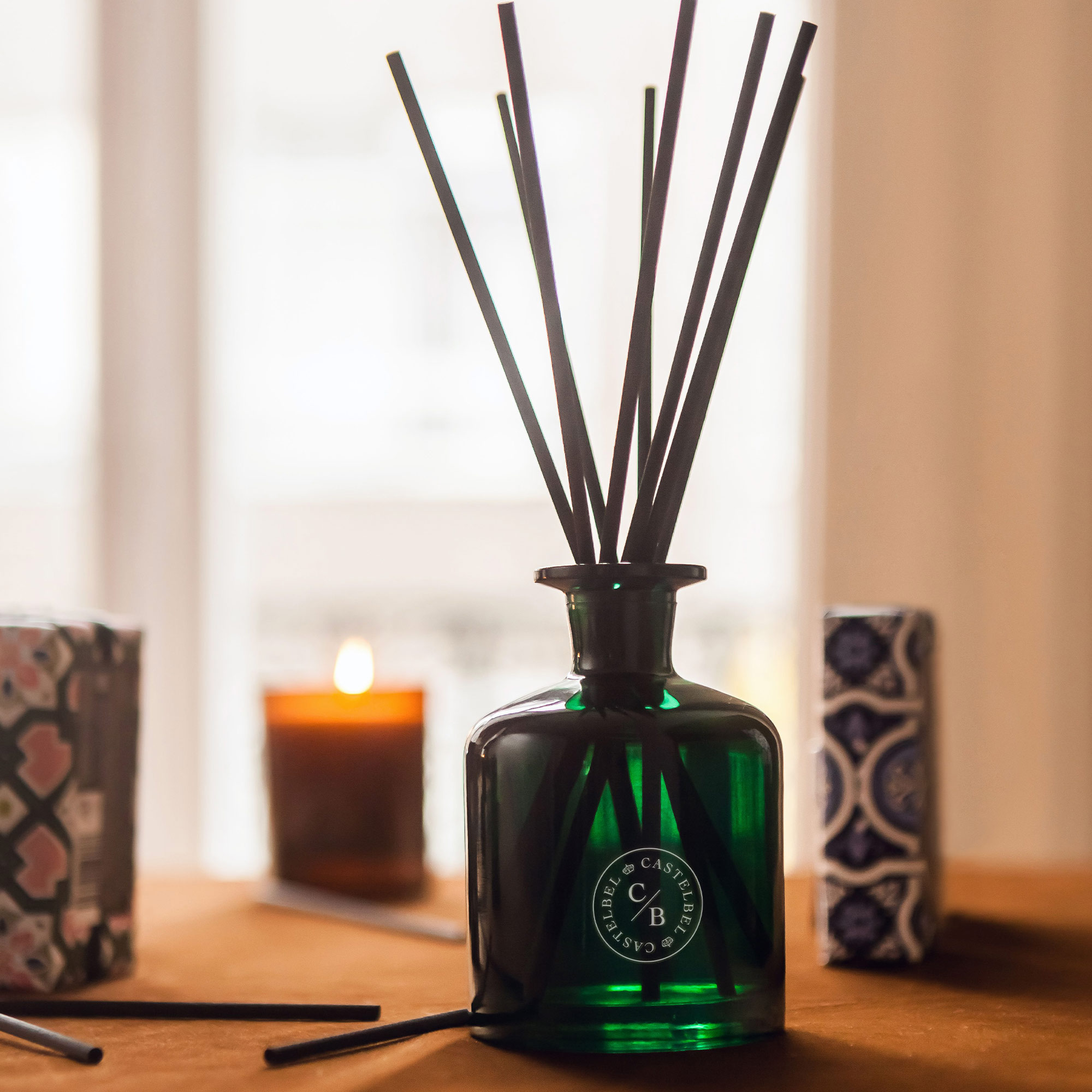 A green glass diffuser with black reeds on a table, surrounded by decorative boxes and a lit candle in the background.