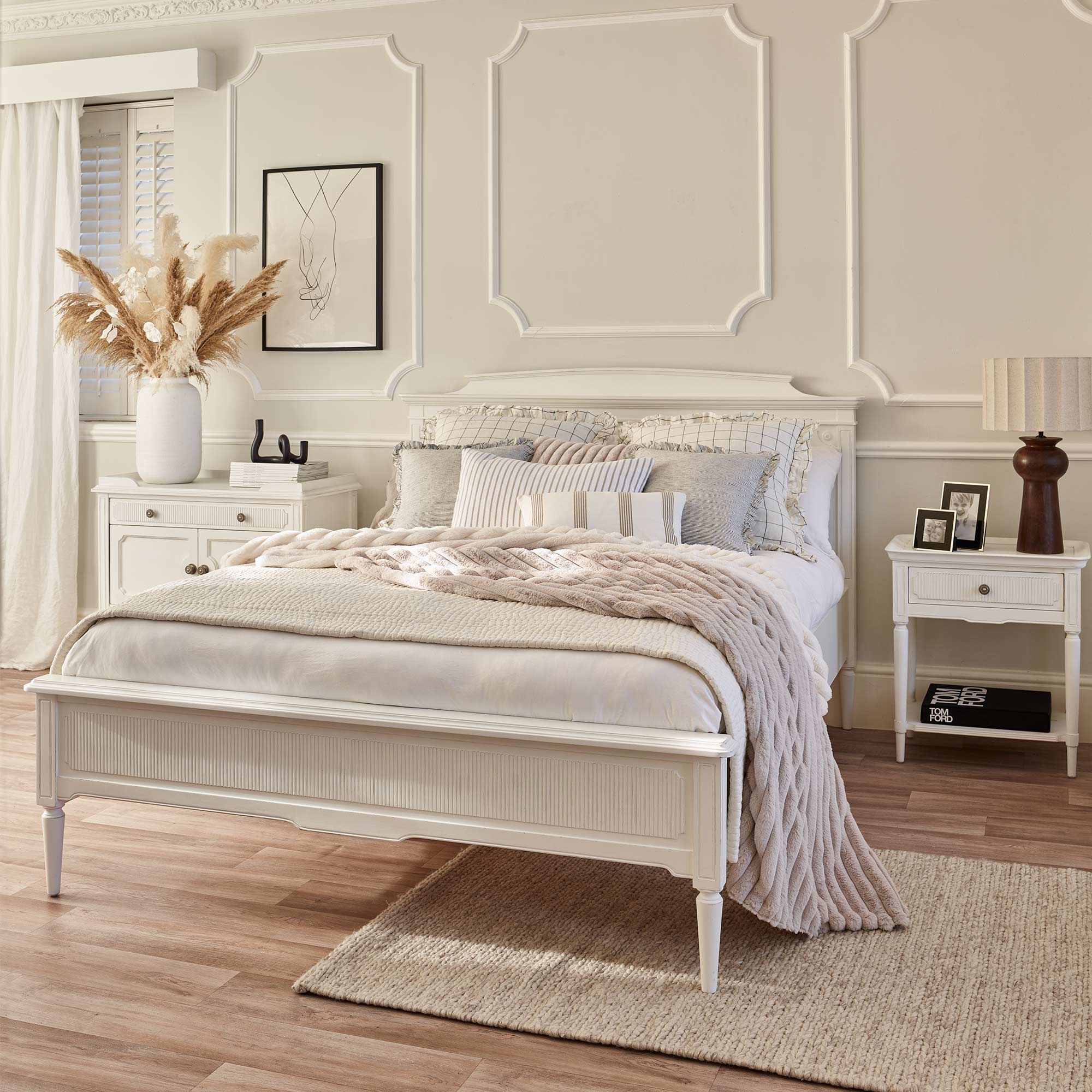 Elegant bedroom with a white bed, beige knitted throw, pampas grass arrangement, framed art, and wooden nightstands on a light wood floor.