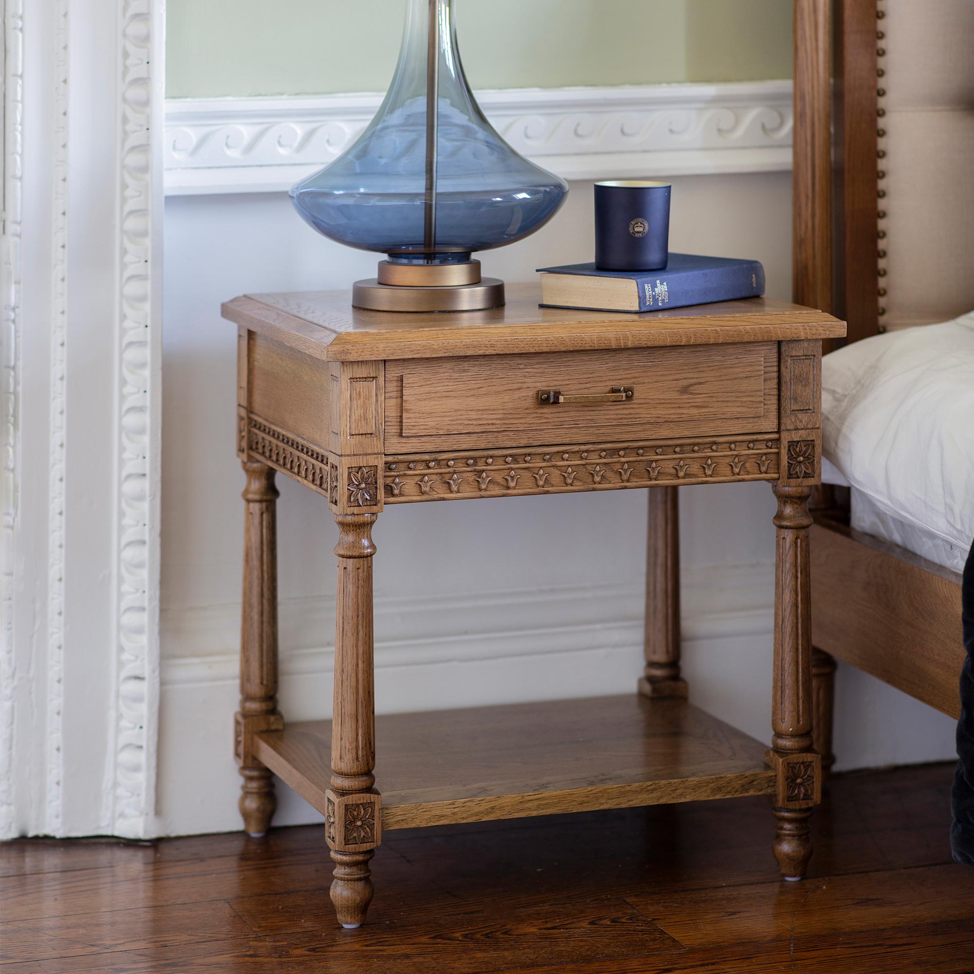Ornate wooden nightstand with a drawer, holding a blue glass lamp, two books, and a small blue container, next to a bed.