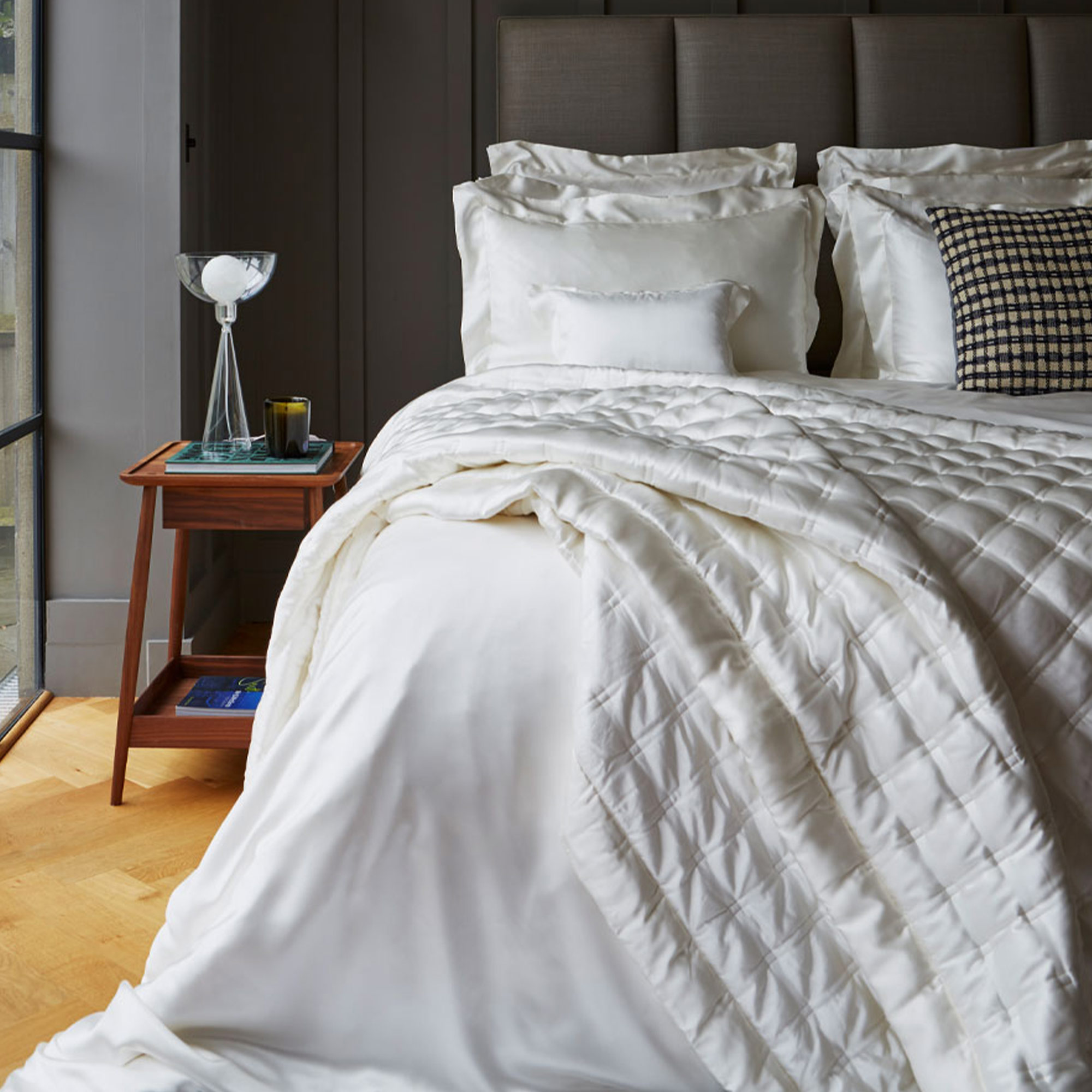 Luxurious bed with white satin sheets and pillows, a checkered cushion, and a wooden side table with a lamp and decor items.