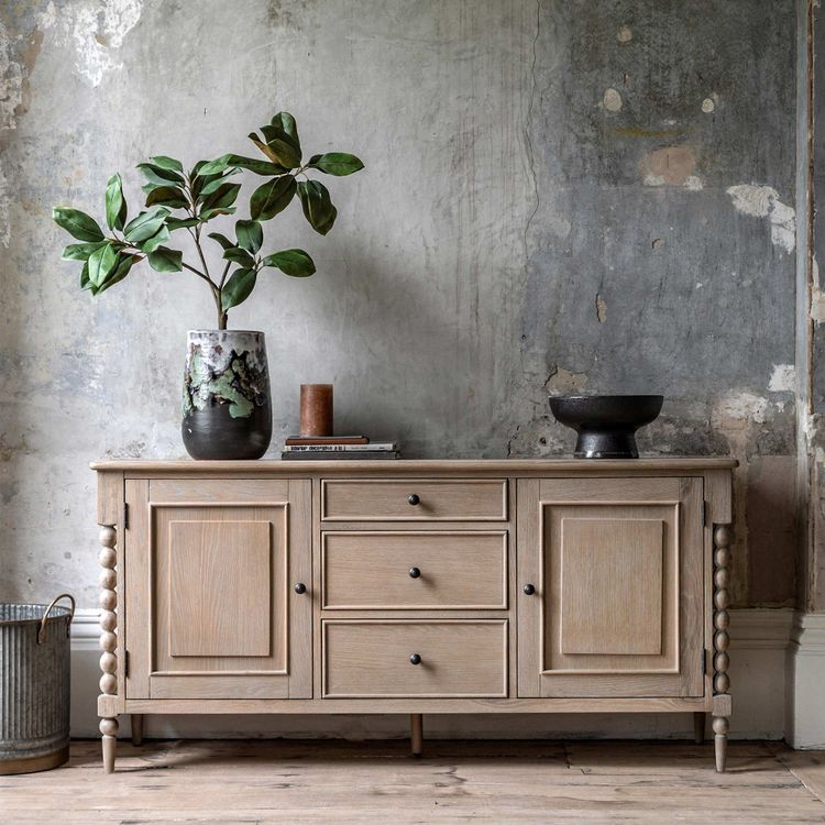 Wooden sideboard with drawers, a plant in a vase, a small brown container, and a dark bowl, set against a rustic, textured wall.