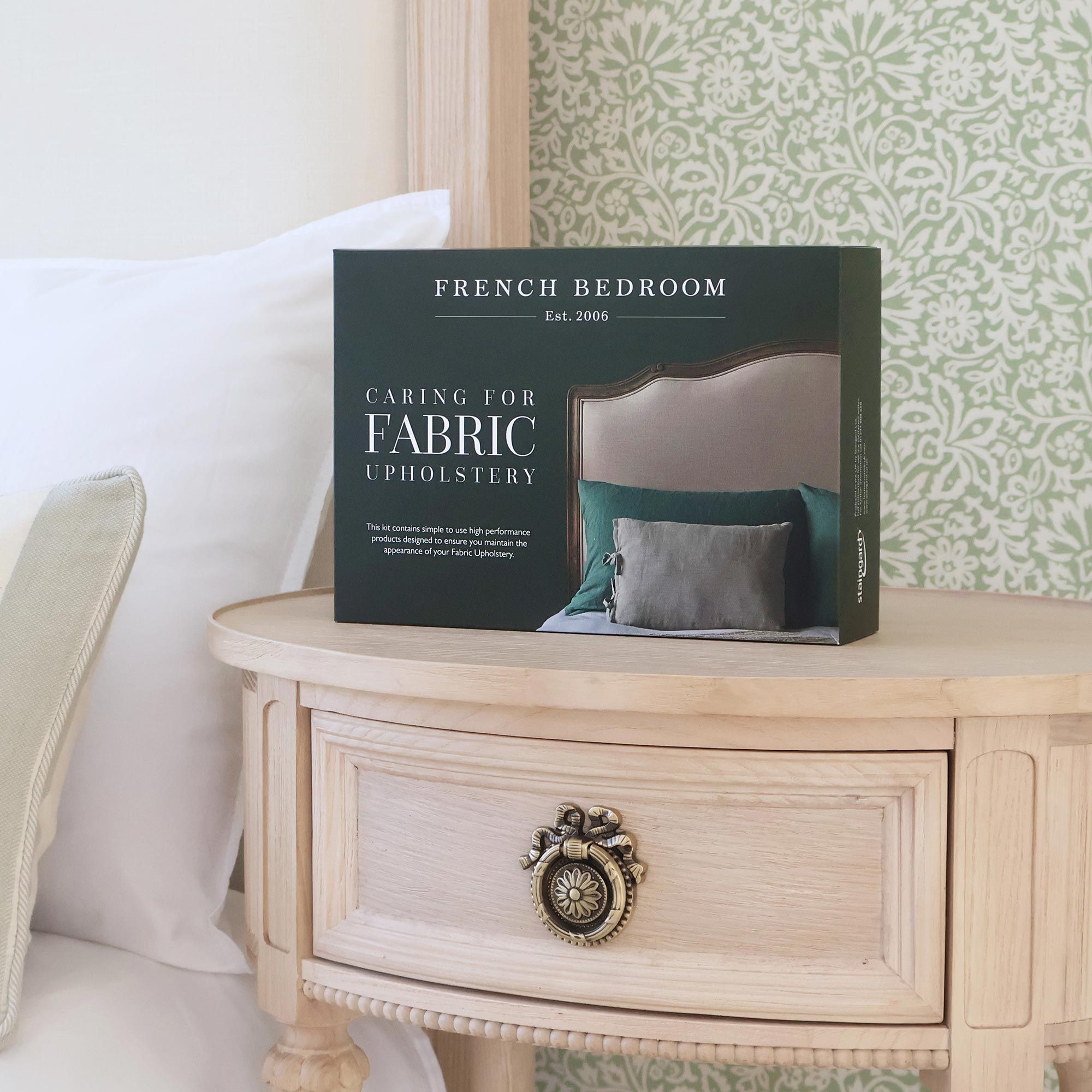 Box labeled "Caring for Fabric Upholstery" on a wooden nightstand with a decorative knob, beside a bed with patterned wallpaper.
