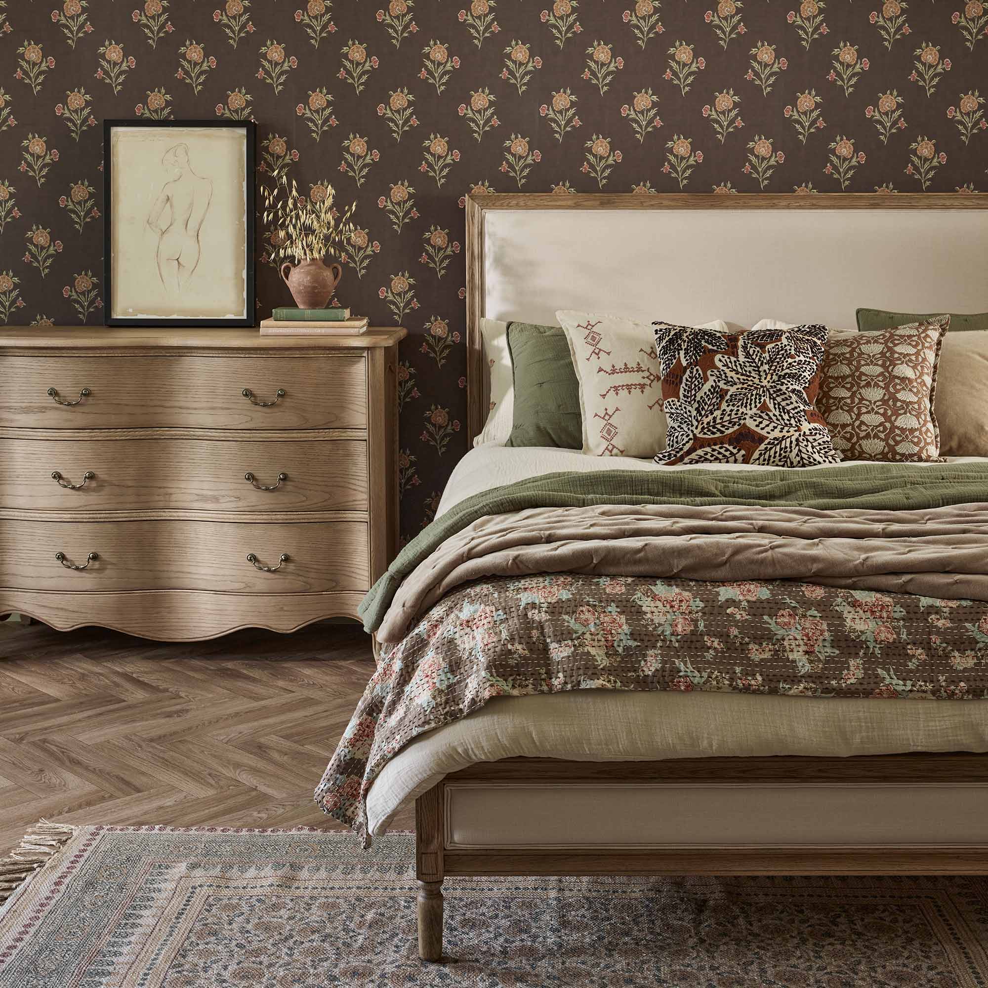 Cozy bedroom with a floral-patterned bed, wooden dresser, framed art, and floral wallpaper. Neutral and green tones create a warm atmosphere.