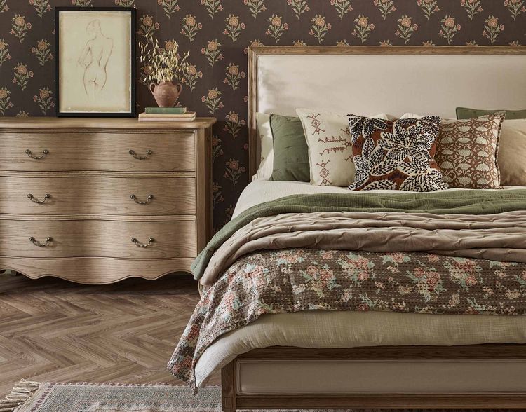 Cozy bedroom with a floral-patterned bed, wooden dresser, framed art, and floral wallpaper. Neutral and green tones create a warm atmosphere.