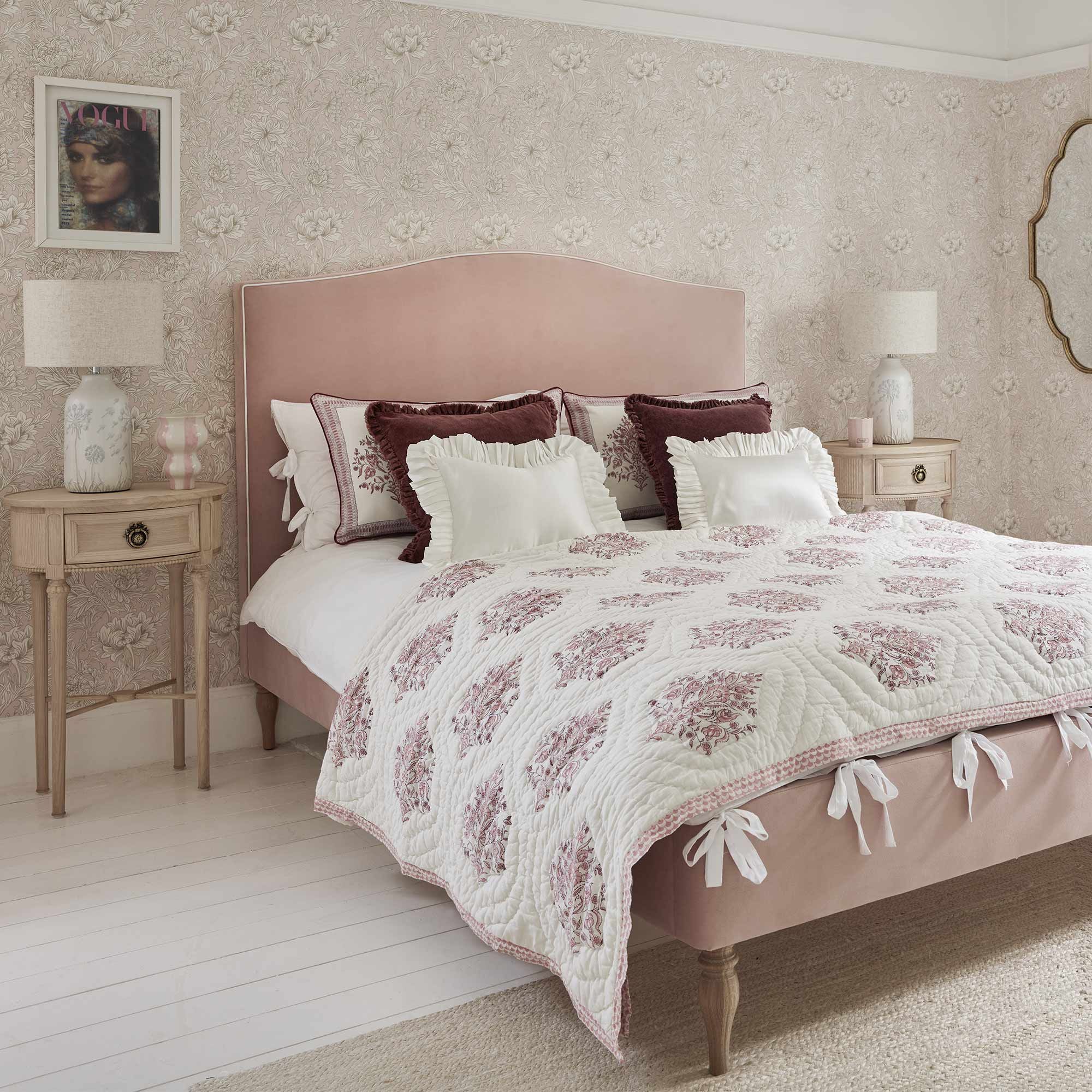 Cozy bedroom with a pink upholstered bed, floral quilt, decorative pillows, vintage wallpaper, and elegant wooden nightstands with lamps.