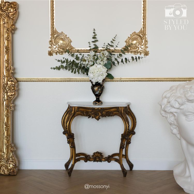 Elegant room with ornate gold mirror, decorative table with floral arrangement, and a classical bust. Parquet flooring adds warmth.