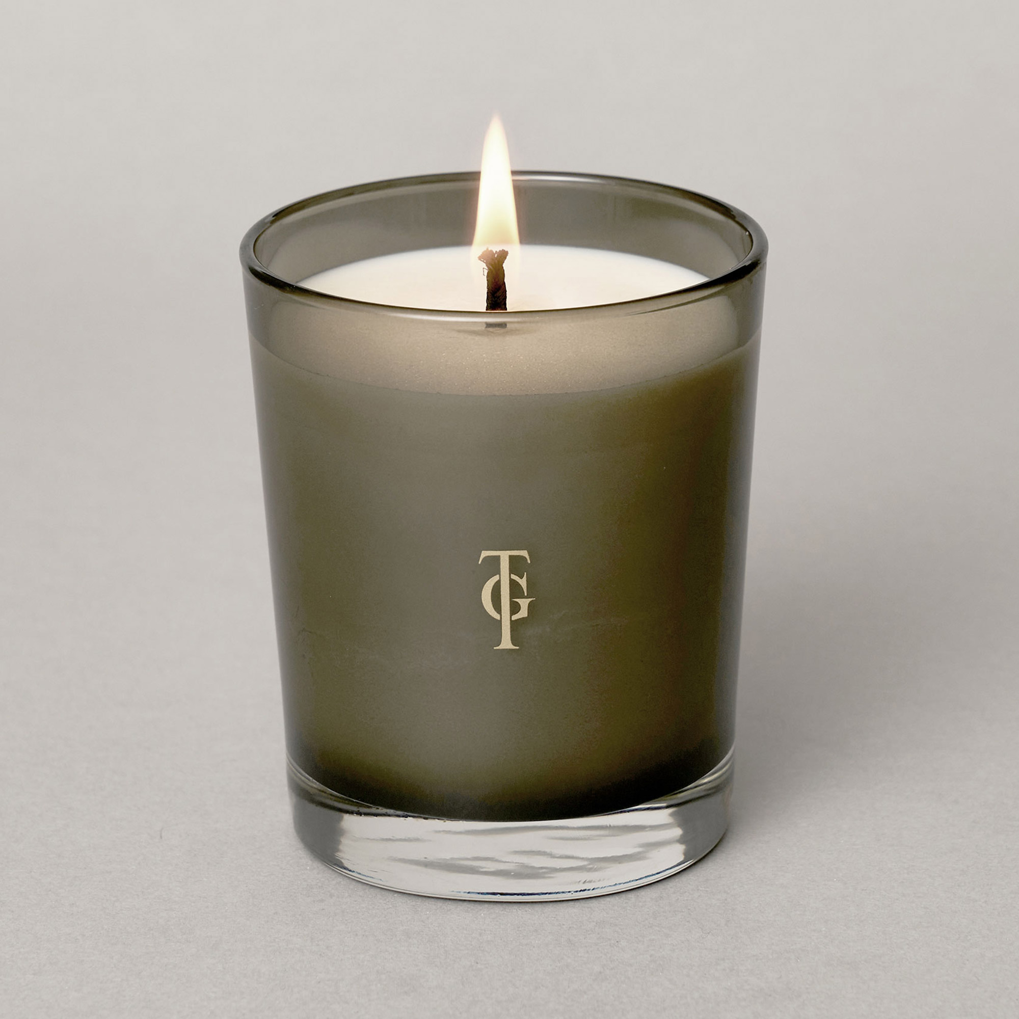 Lit candle in a dark glass holder with a gold monogram on the front, set against a plain gray background.