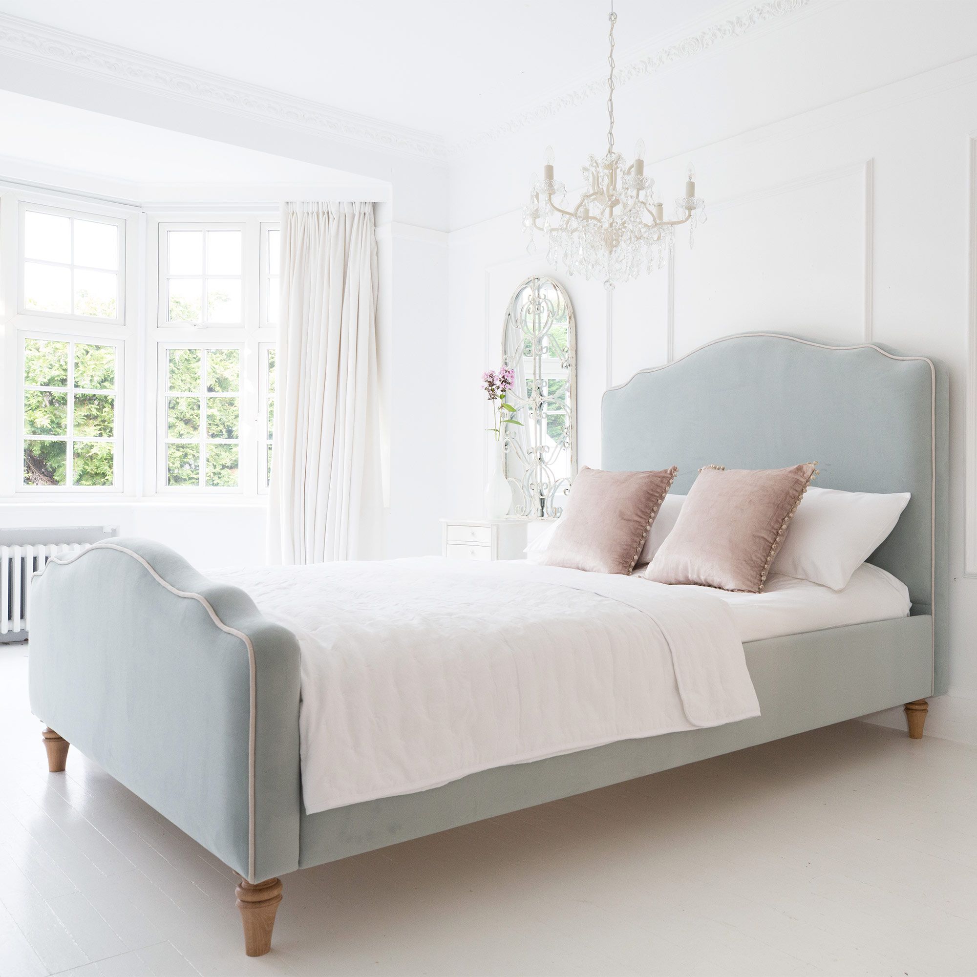 Elegant bedroom with a light blue upholstered bed, white bedding, pink pillows, a chandelier, and a large window with curtains.