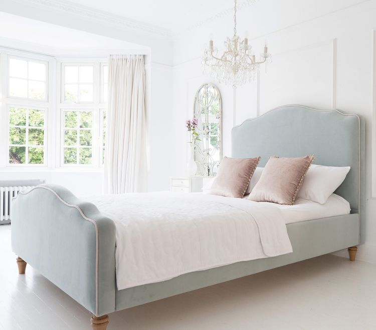 Elegant bedroom with a light blue upholstered bed, white bedding, pink pillows, a chandelier, and a large window with curtains.