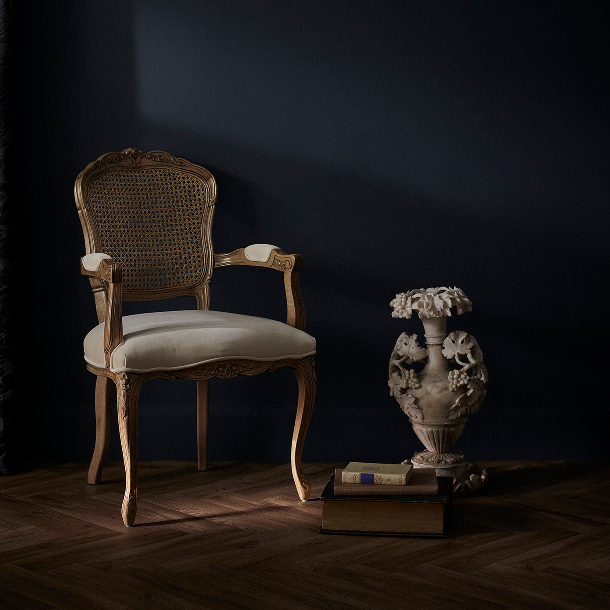 Elegant wooden chair with woven backrest next to a decorative vase and books on a dark wooden floor against a dark wall