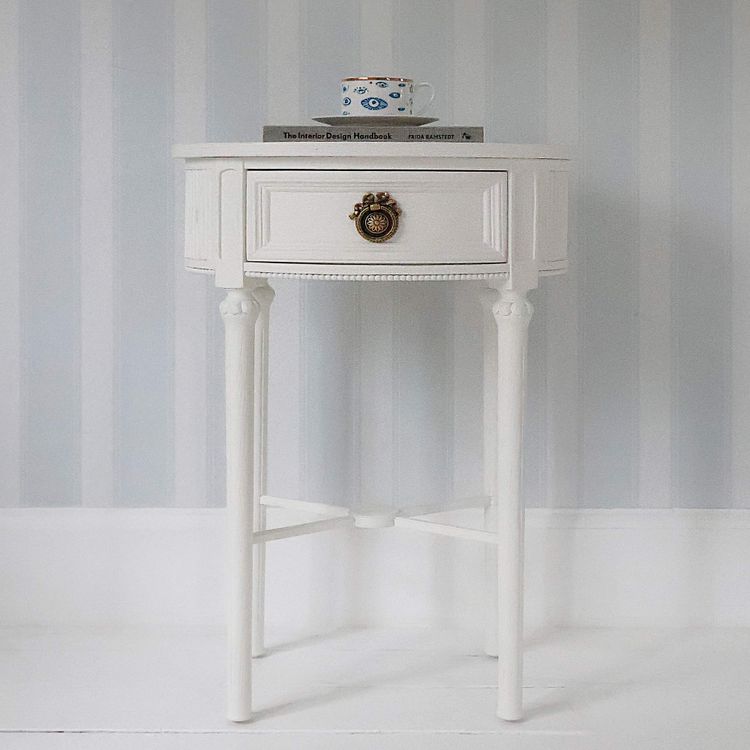 A small white round table with a drawer and ornate handle, holding a mug and two books, set against a striped gray and white wall.