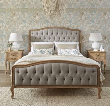 Elegant bedroom with a tufted bed, floral wallpaper, two wooden nightstands, white lamps, and a decorative chandelier above.