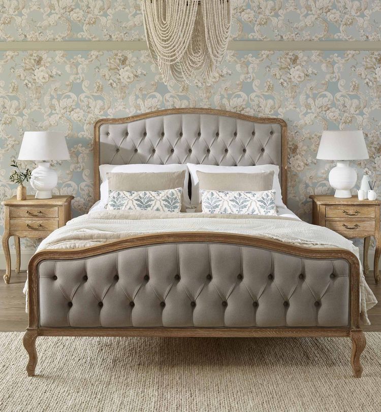 Elegant bedroom with a tufted bed, floral wallpaper, two wooden nightstands, white lamps, and a decorative chandelier above.