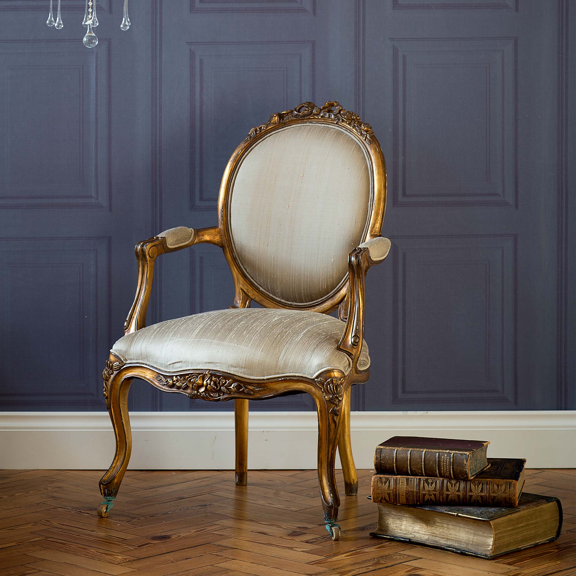 Elegant vintage chair with ornate wooden frame and beige upholstery, set against a paneled gray wall, beside a stack of antique books on wooden floor.