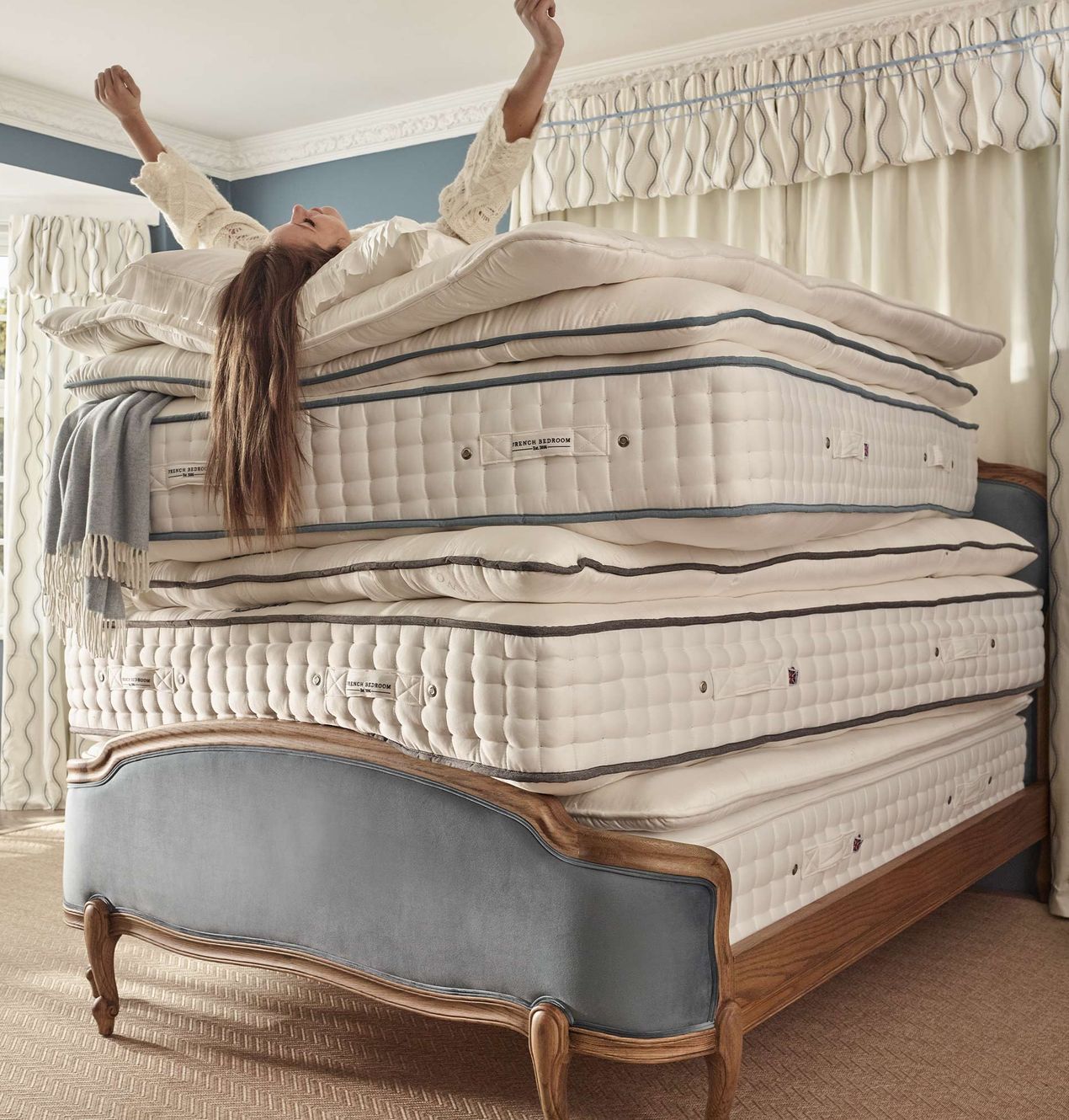 Woman joyfully stretches on a stack of five luxury mattresses on an upholstered French bed.