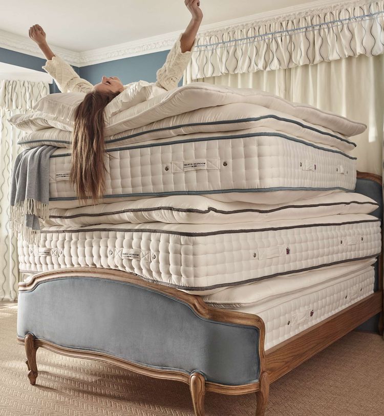 Woman joyfully stretches on a stack of five luxury mattresses on an upholstered French bed.