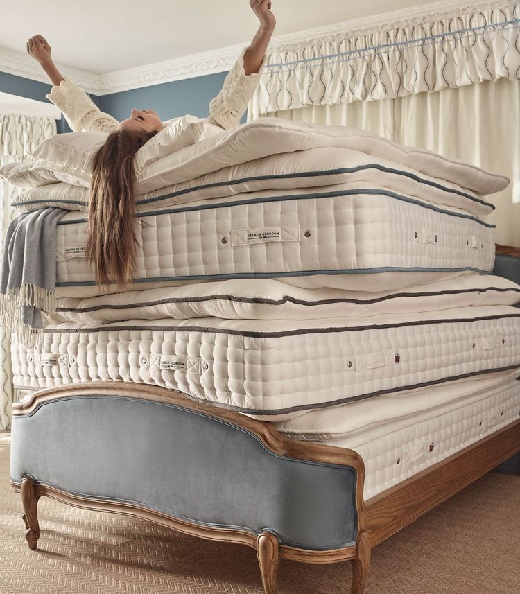 Woman joyfully stretches on a stack of five luxury mattresses on an upholstered French bed.