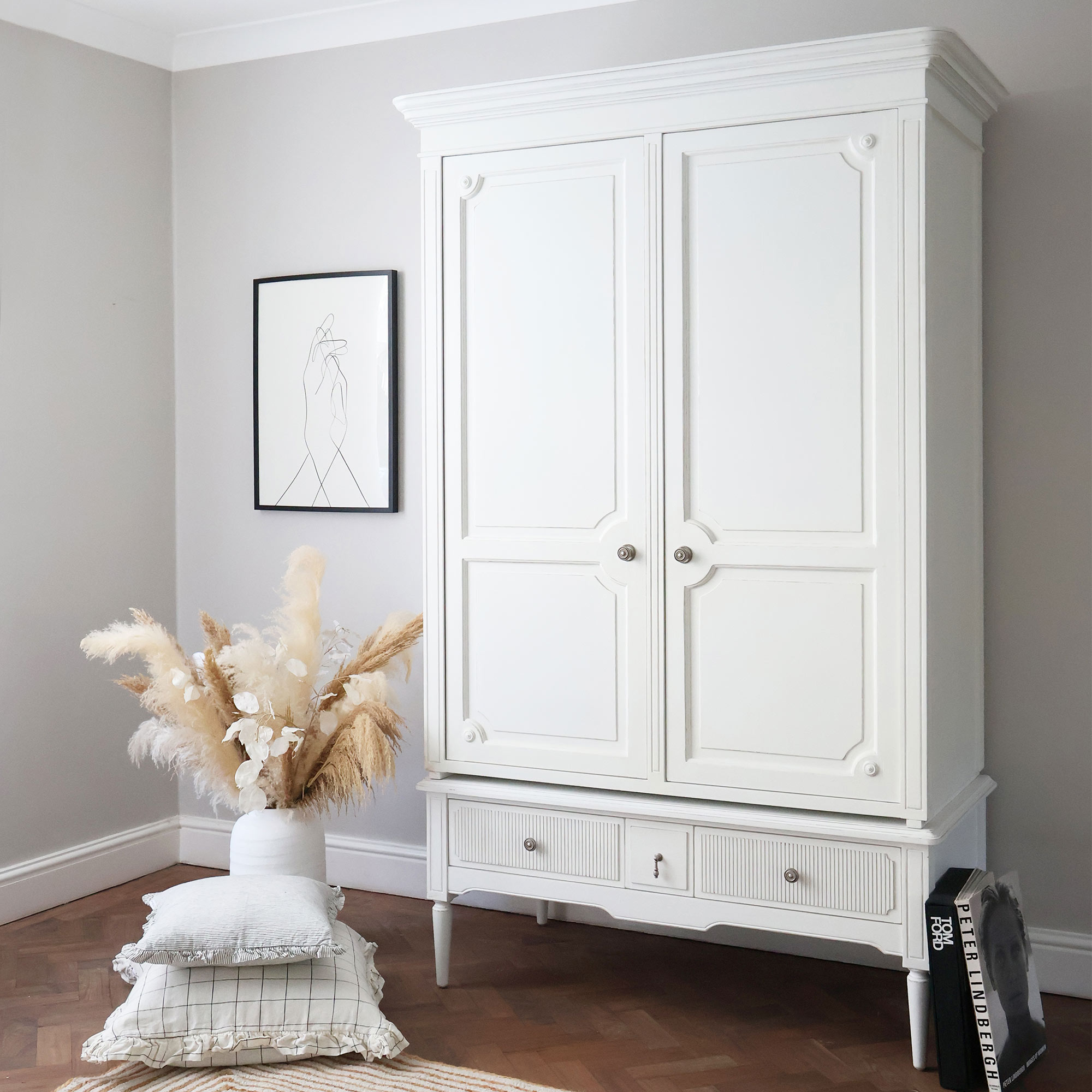 White wardrobe in a minimalist room with a line art portrait, pampas grass in a vase, and stacked pillows on the floor.