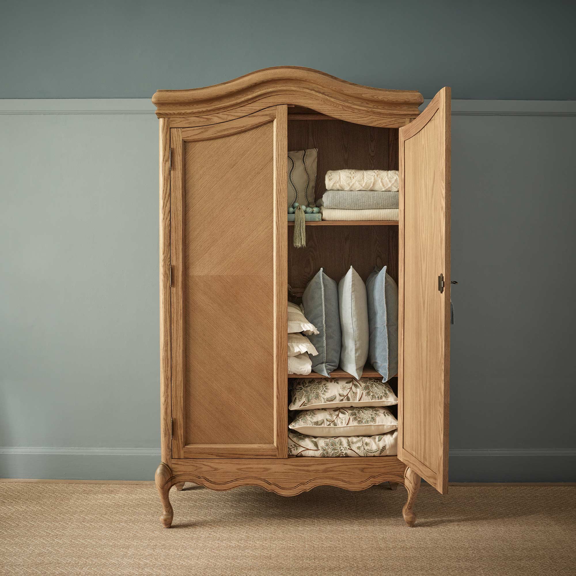 Solid wooden wardrobe with an open door revealing folded blankets, cushions, and pillows inside, set against a muted blue wall.