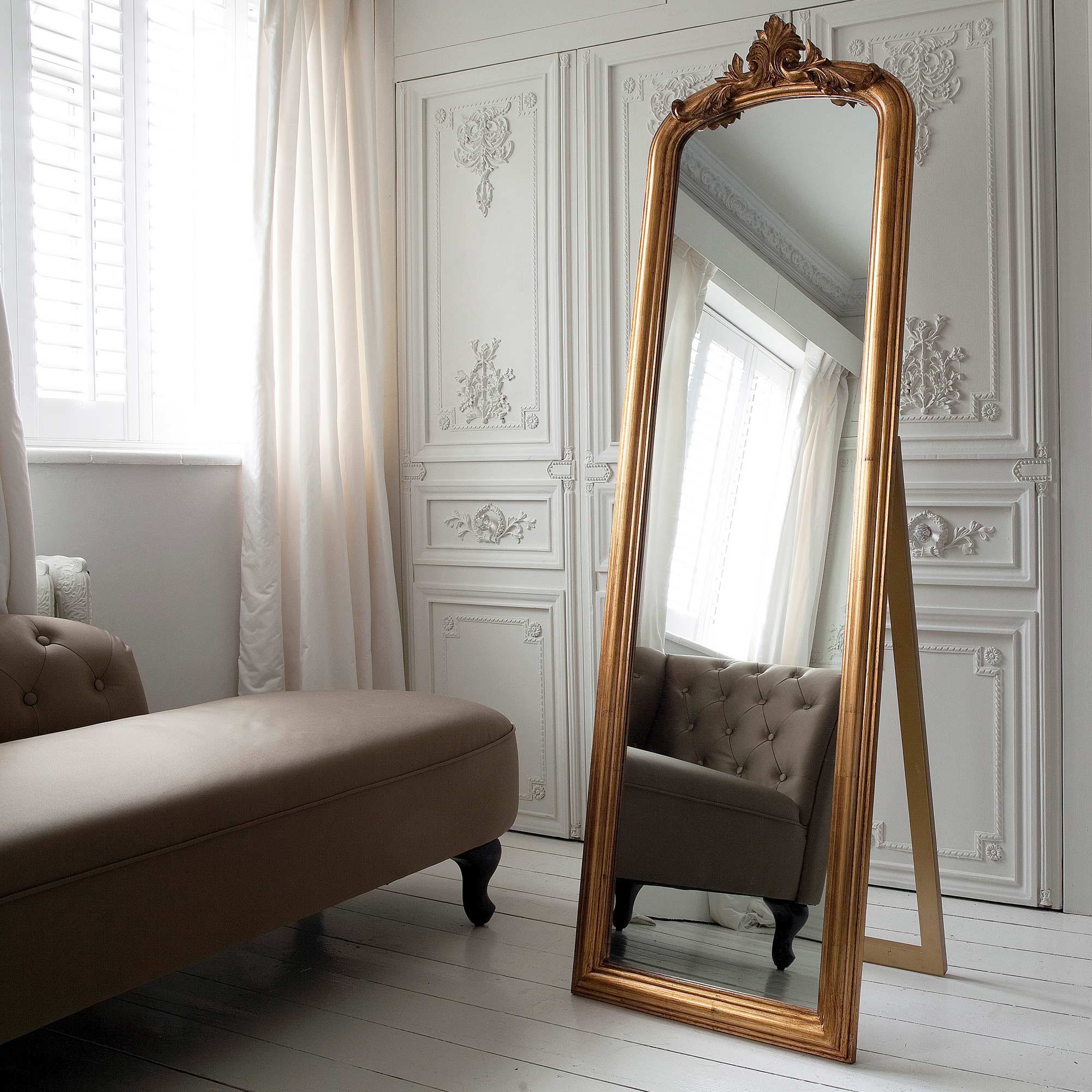 Elegant room with a tall ornate gold mirror reflecting a tufted sofa, white paneled walls, and sheer curtains.