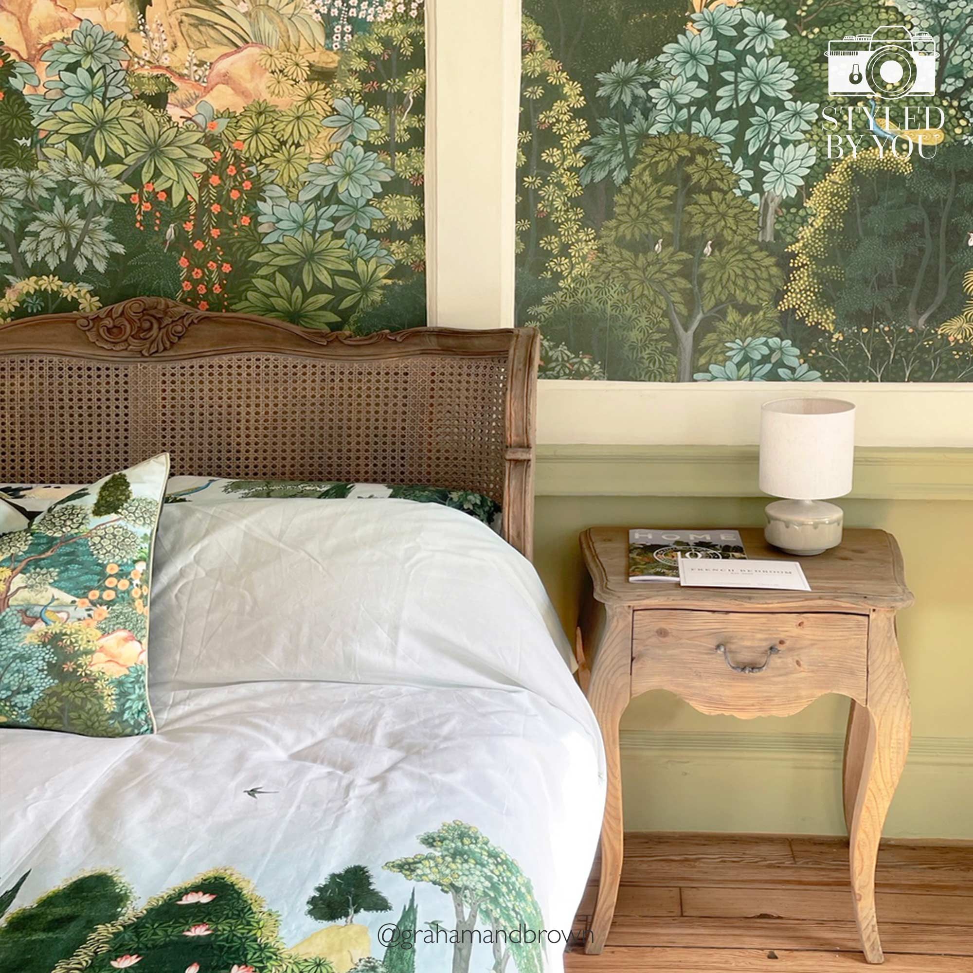Cozy bedroom with a wooden bed, nature-themed bedding, and a vintage nightstand. Lush, green botanical wallpaper in the background.