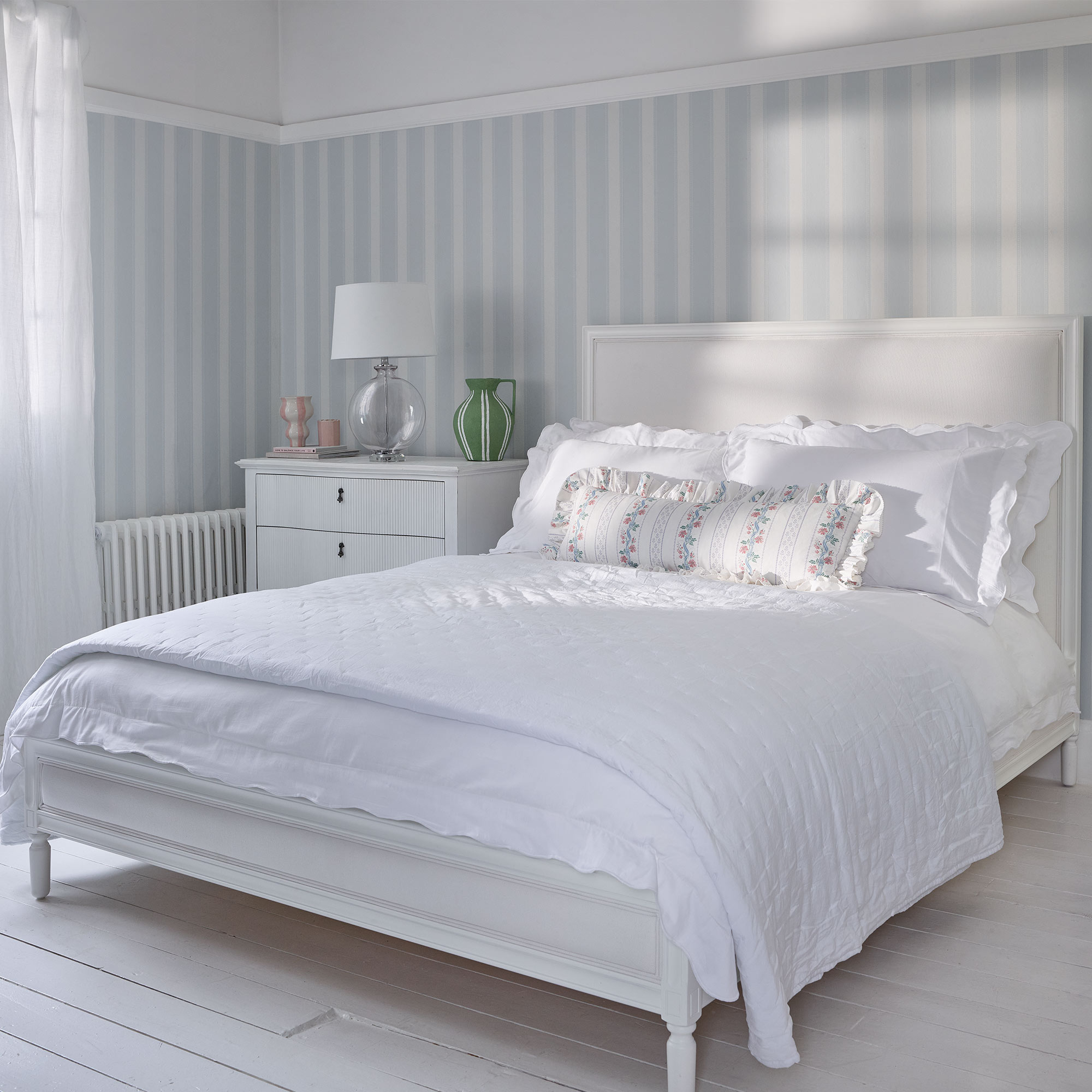 Bright bedroom with a white bed, floral pillows, striped wallpaper, and a white dresser. Natural light filters through sheer curtains.
