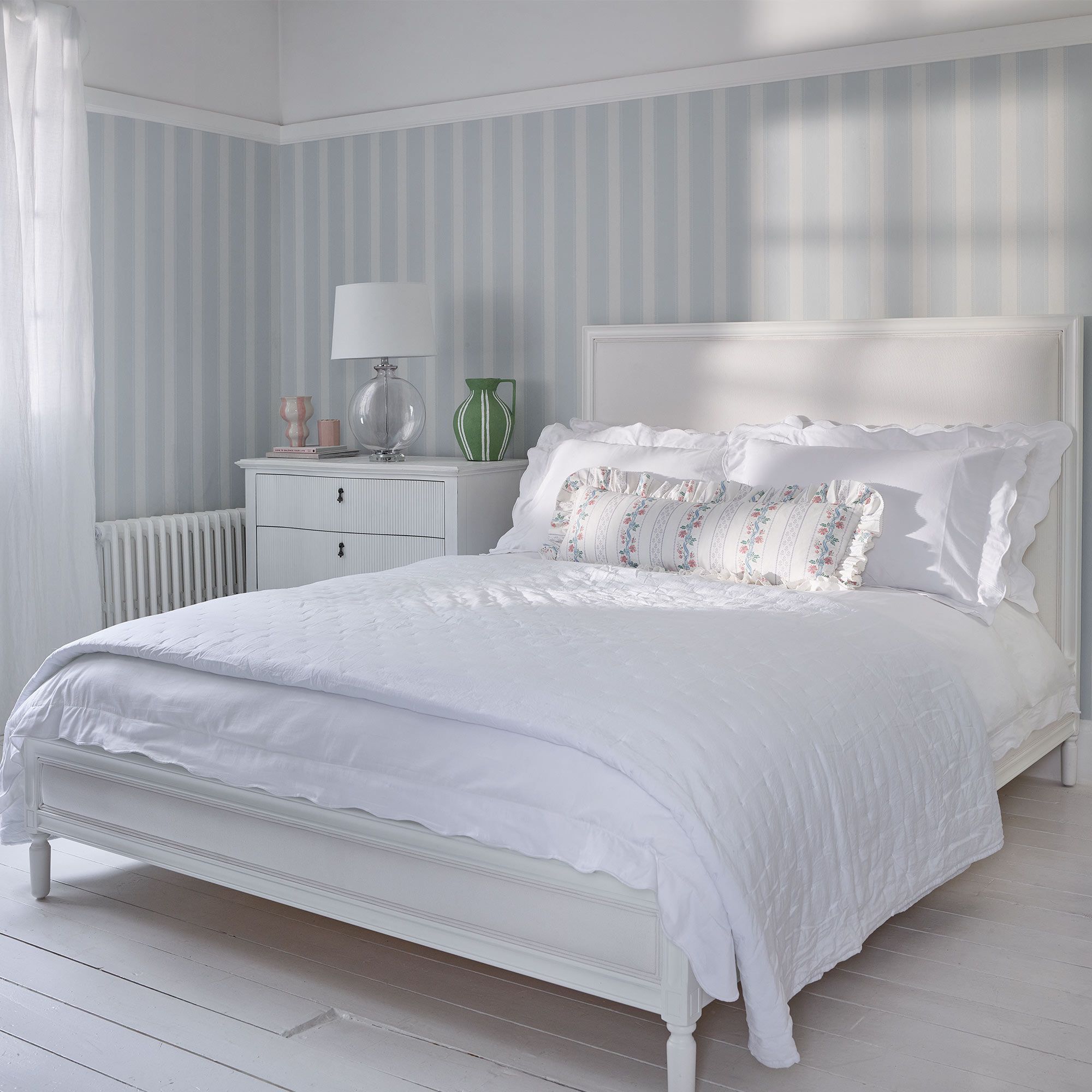 Bright bedroom with a white bed, floral pillows, striped wallpaper, and a white dresser. Natural light filters through sheer curtains.