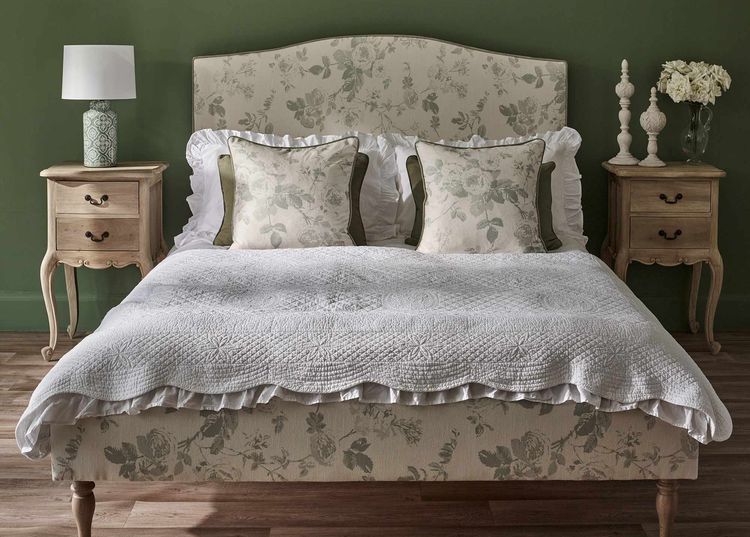 Elegant bedroom with a floral-patterned bed, white quilt, and two pillows. Wooden nightstands with lamps flank the bed against a green wall.