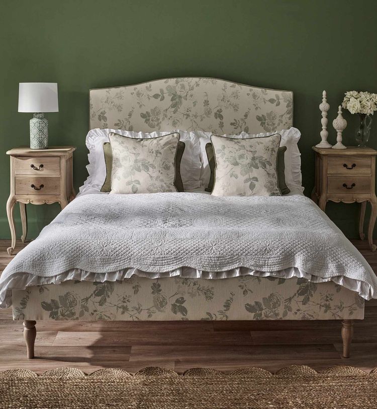 Elegant bedroom with a floral-patterned bed, white quilt, and two pillows. Wooden nightstands with lamps flank the bed against a green wall.