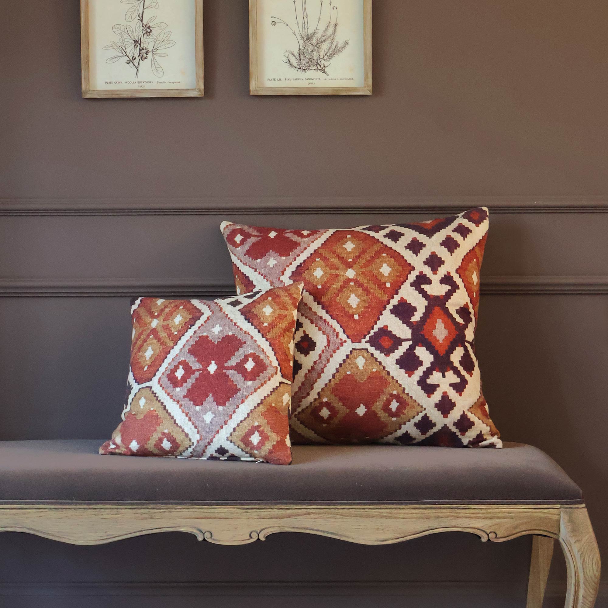 Two patterned cushions with geometric designs on a wooden bench, set against a brown wall with framed botanical sketches.