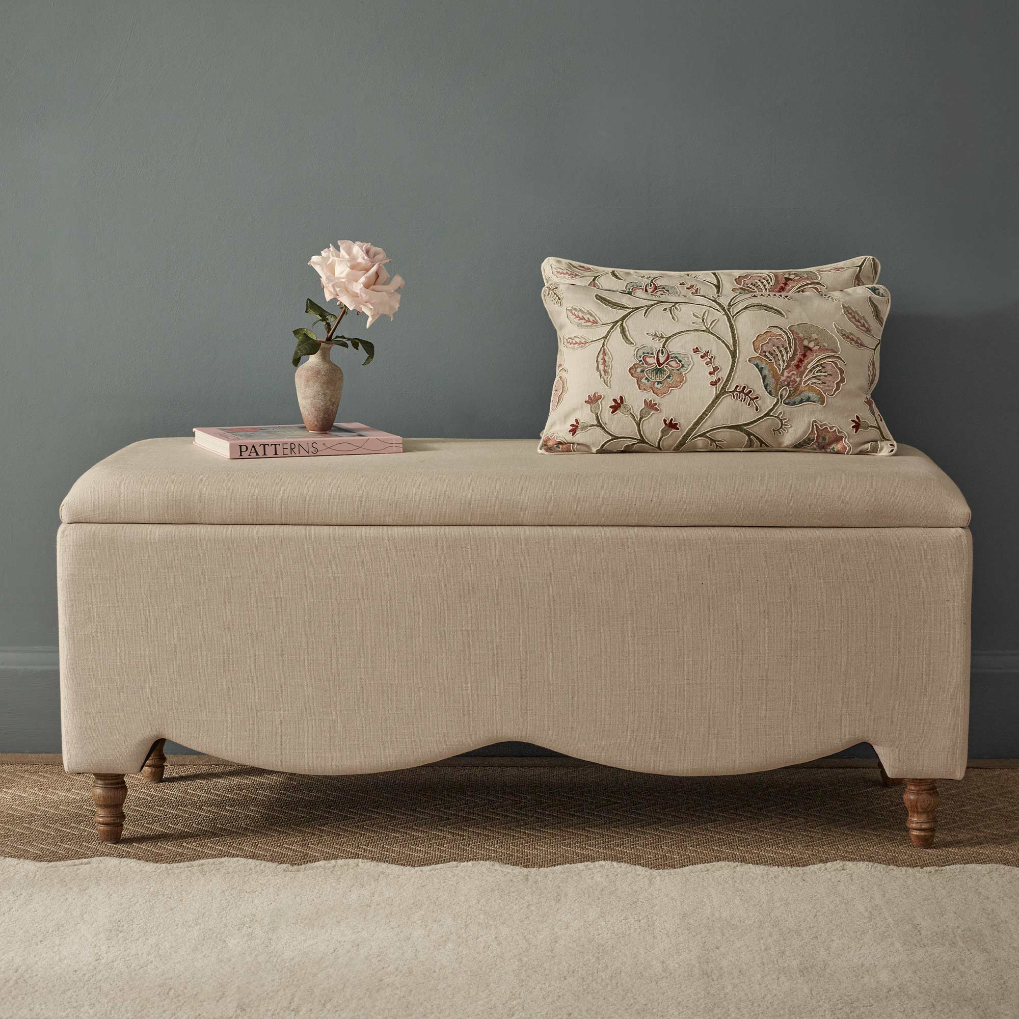 Beige ottoman in linen with a floral pillow, a pink book, and a vase with a pink rose, set against a blue wall on a beige wavy rug.