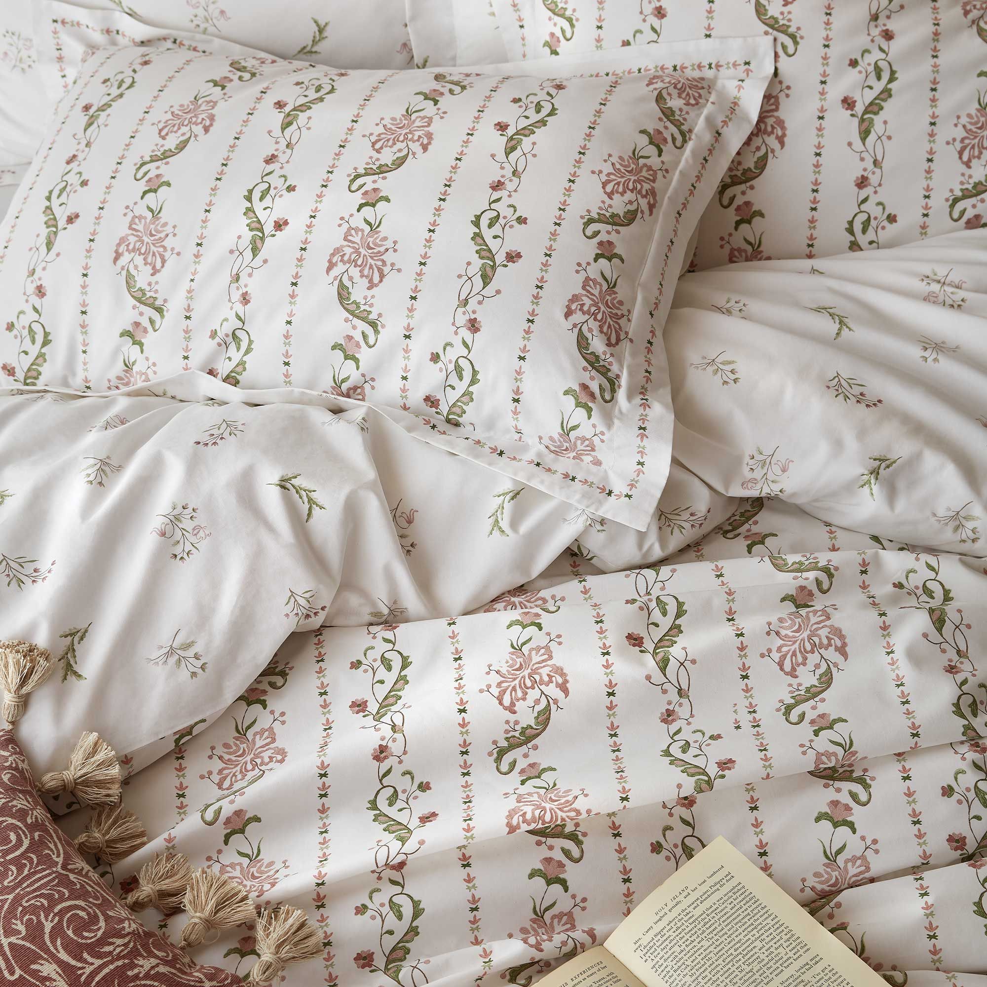 Floral-patterned bedding in pink and green tones with an open book on a bed, featuring intricate designs inspired  by an 1850 gentleman’s ivory silk waistcoat housed in the Royal Ceremonial Dress Collection.