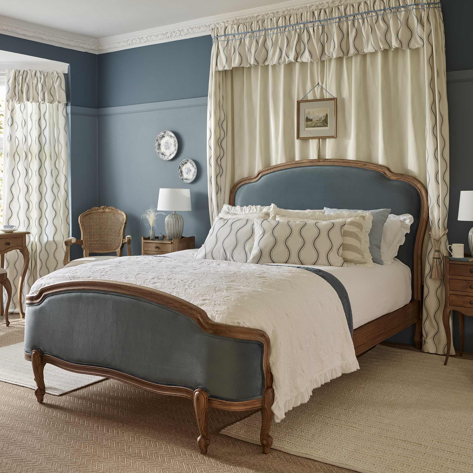 Elegant bedroom with a vintage-style blue upholstered wooden bed, blue and white decor, patterned curtains, and a cozy armchair beside a wooden nightstand.