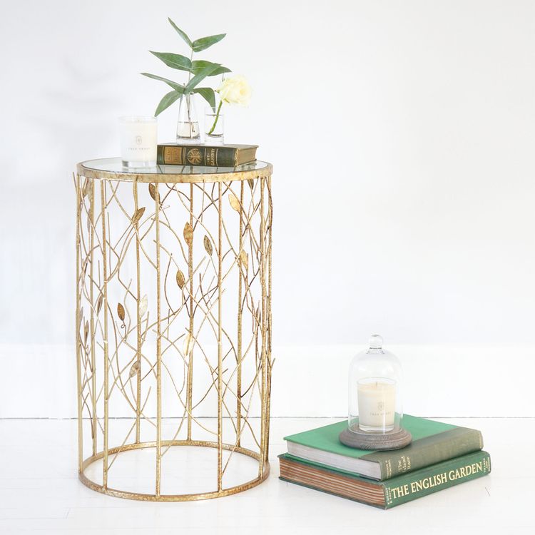 Elegant gold side table with leaf design, topped with books, a candle, and a plant. Nearby, a stack of books with another candle on top.