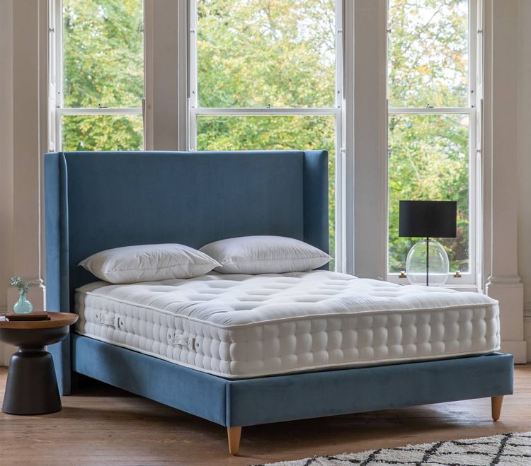 Blue upholstered bed with a white mattress and pillows in a bright room with large windows, a black lamp, and a small round table.