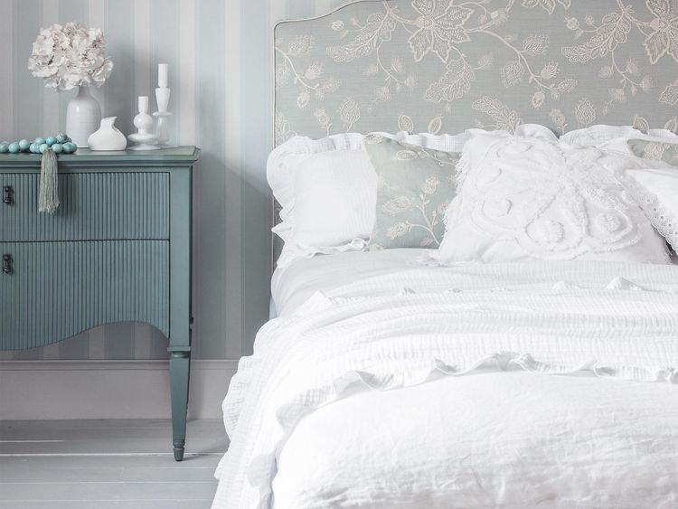 Elegant bedroom with a floral headboard, white bedding, and a teal nightstand. Soft lighting and striped wallpaper create a serene atmosphere.