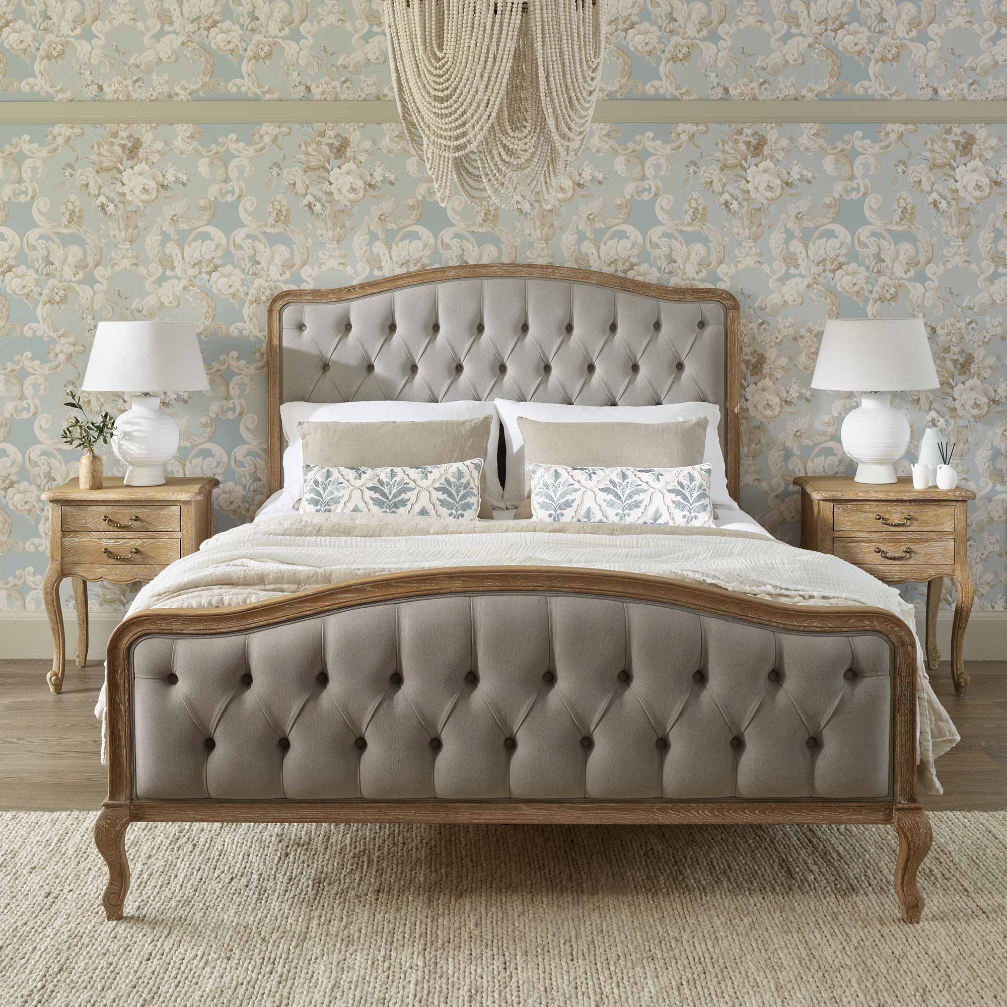 Elegant bedroom with a tufted bed, floral wallpaper, and matching wooden nightstands with lamps. A chandelier hangs above.