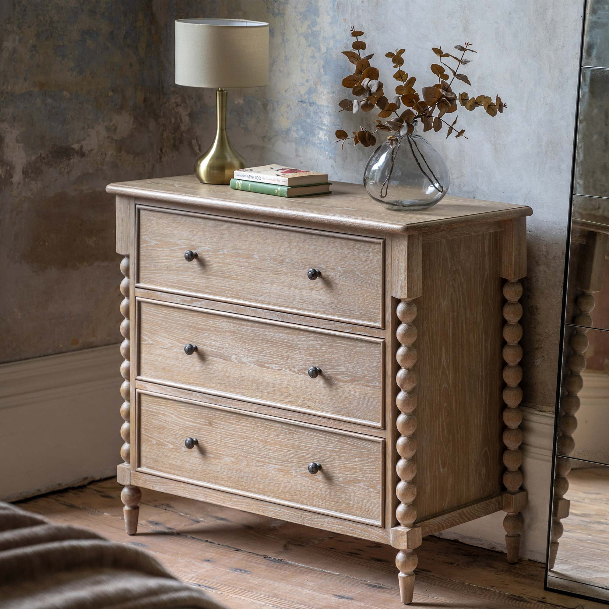 Wooden dresser with three drawers and decorative knobs, topped with a lamp, books, and a vase with dried leaves, beside a large mirror.