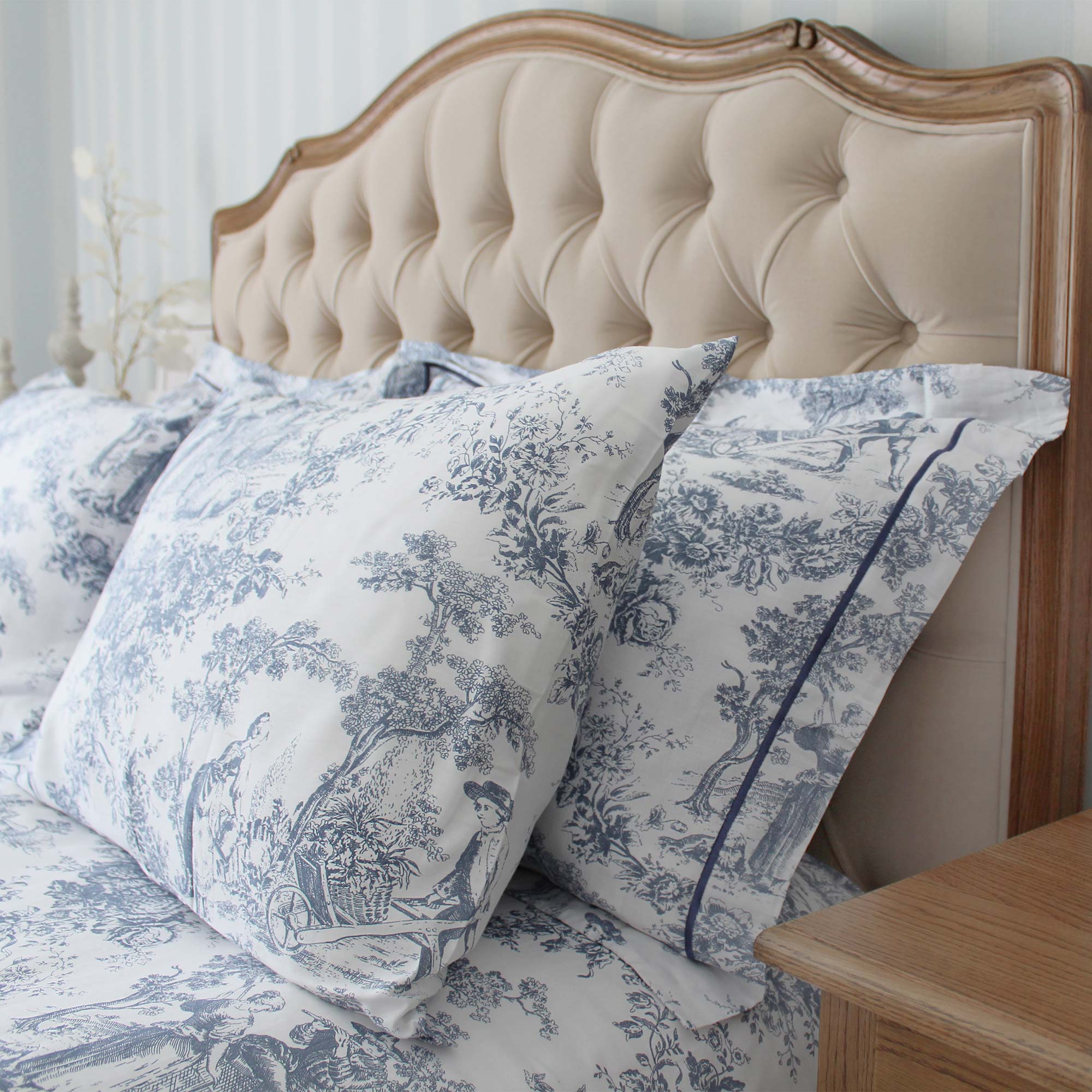 A bed with a tufted beige headboard, featuring pillows and sheets with a blue and white toile pattern, next to a wooden nightstand.