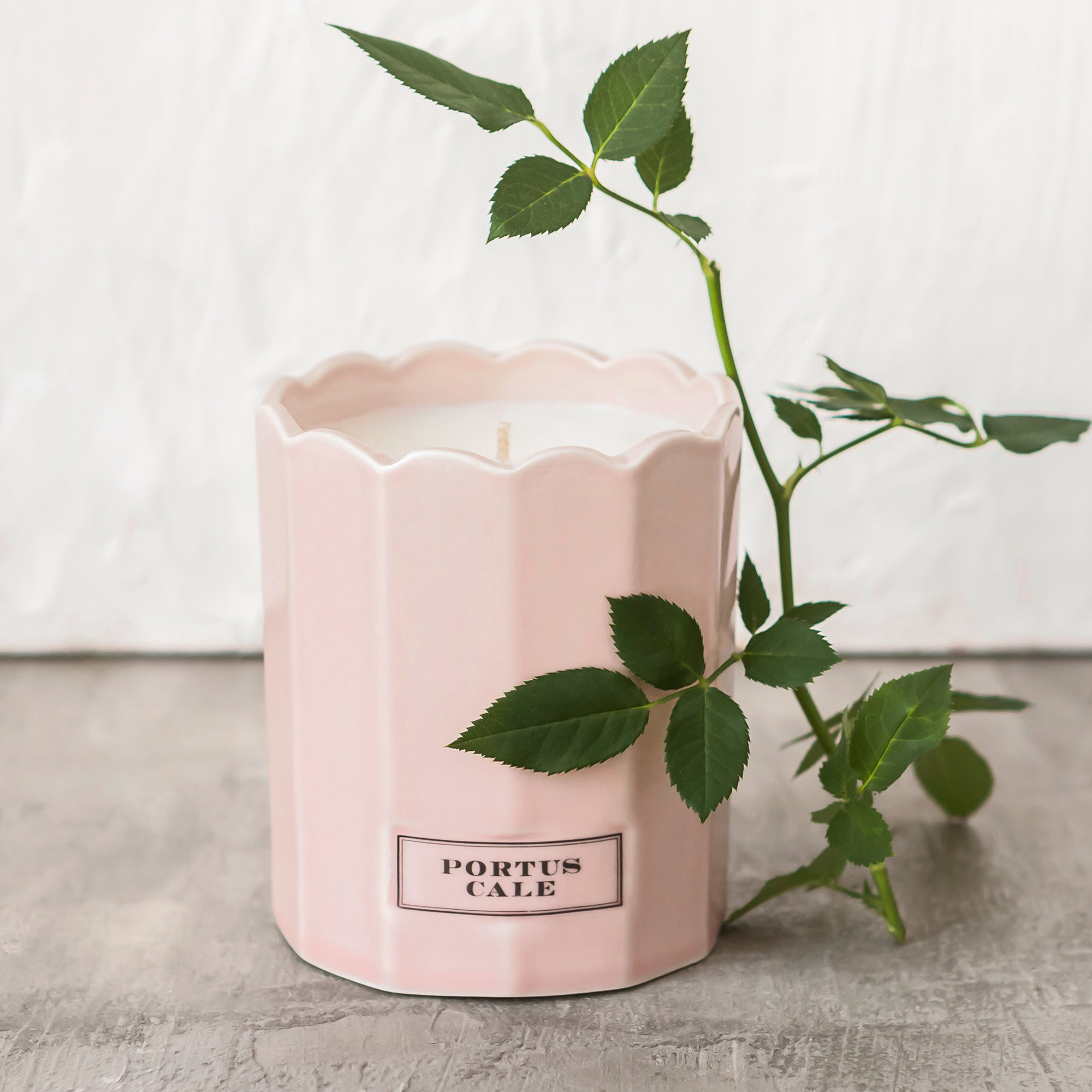 A pink candle in a fluted ceramic holder with "Portus Cale" label, adorned with a sprig of green leaves, set on a gray surface.