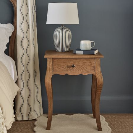 Wooden bedside table with a striped lamp, mug, and book against a dark wall.