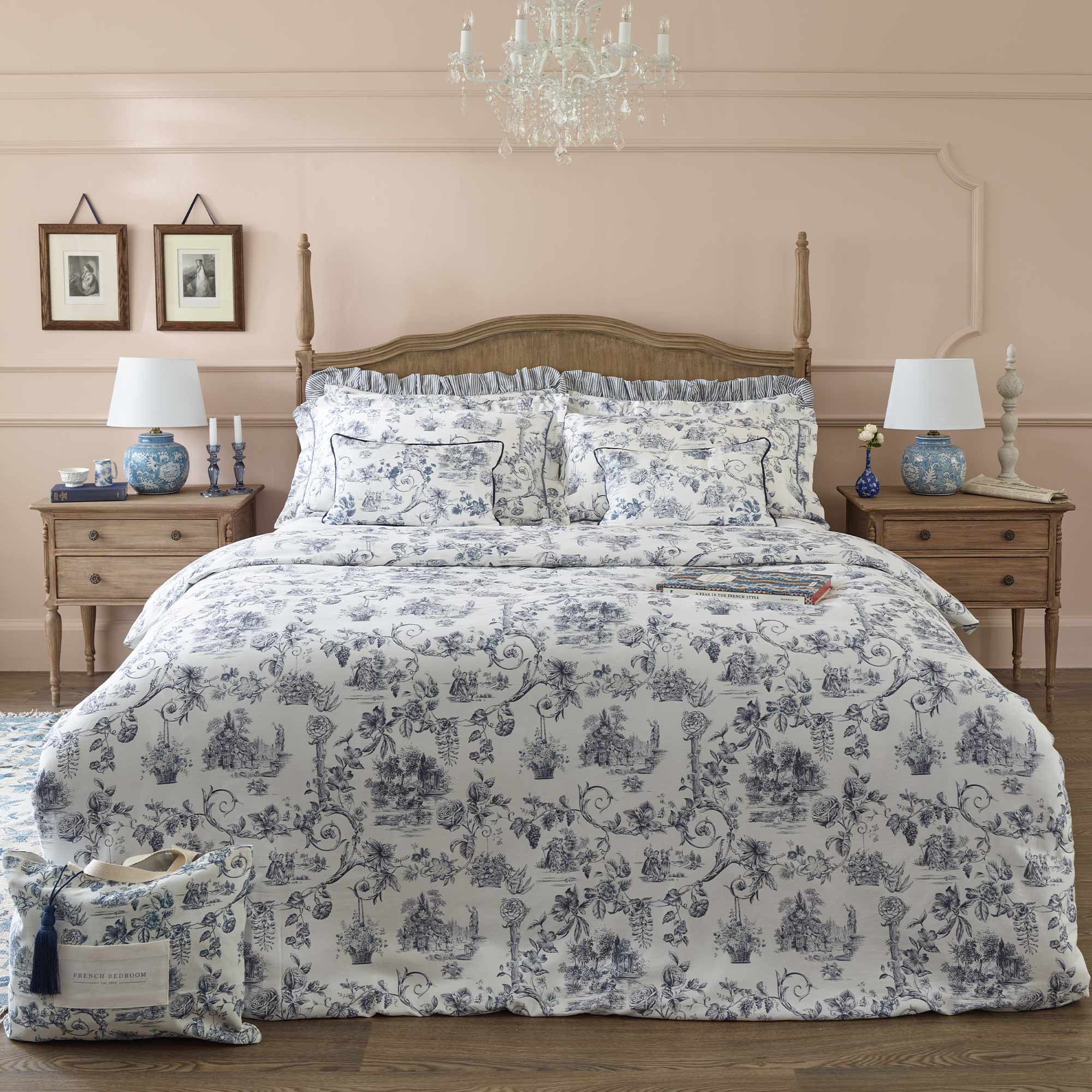Elegant bedroom with a wooden bed, floral-patterned bedding, blue lamps, and framed art. Soft pink walls and a chandelier complete the cozy setting.