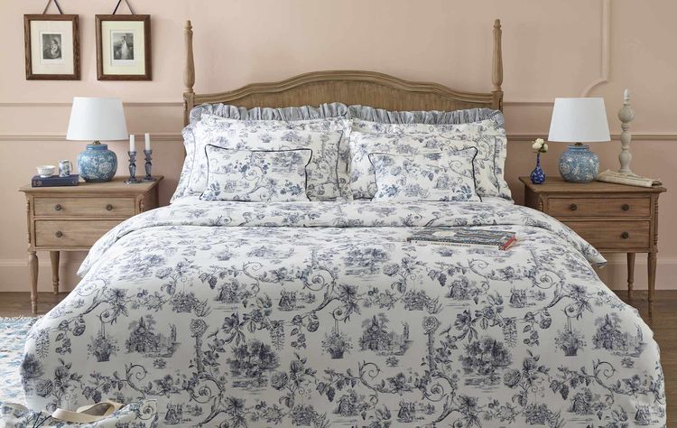 Elegant bedroom with a wooden bed, floral-patterned bedding, blue lamps, and framed art. Soft pink walls and a chandelier complete the cozy setting.