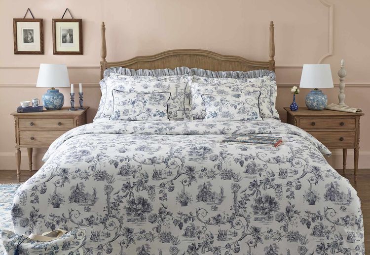 Elegant bedroom with a wooden bed, floral-patterned bedding, blue lamps, and framed art. Soft pink walls and a chandelier complete the cozy setting.