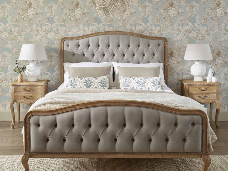 Elegant bedroom with a tufted bed, floral wallpaper, wooden nightstands, white lamps, and a decorative chandelier.
