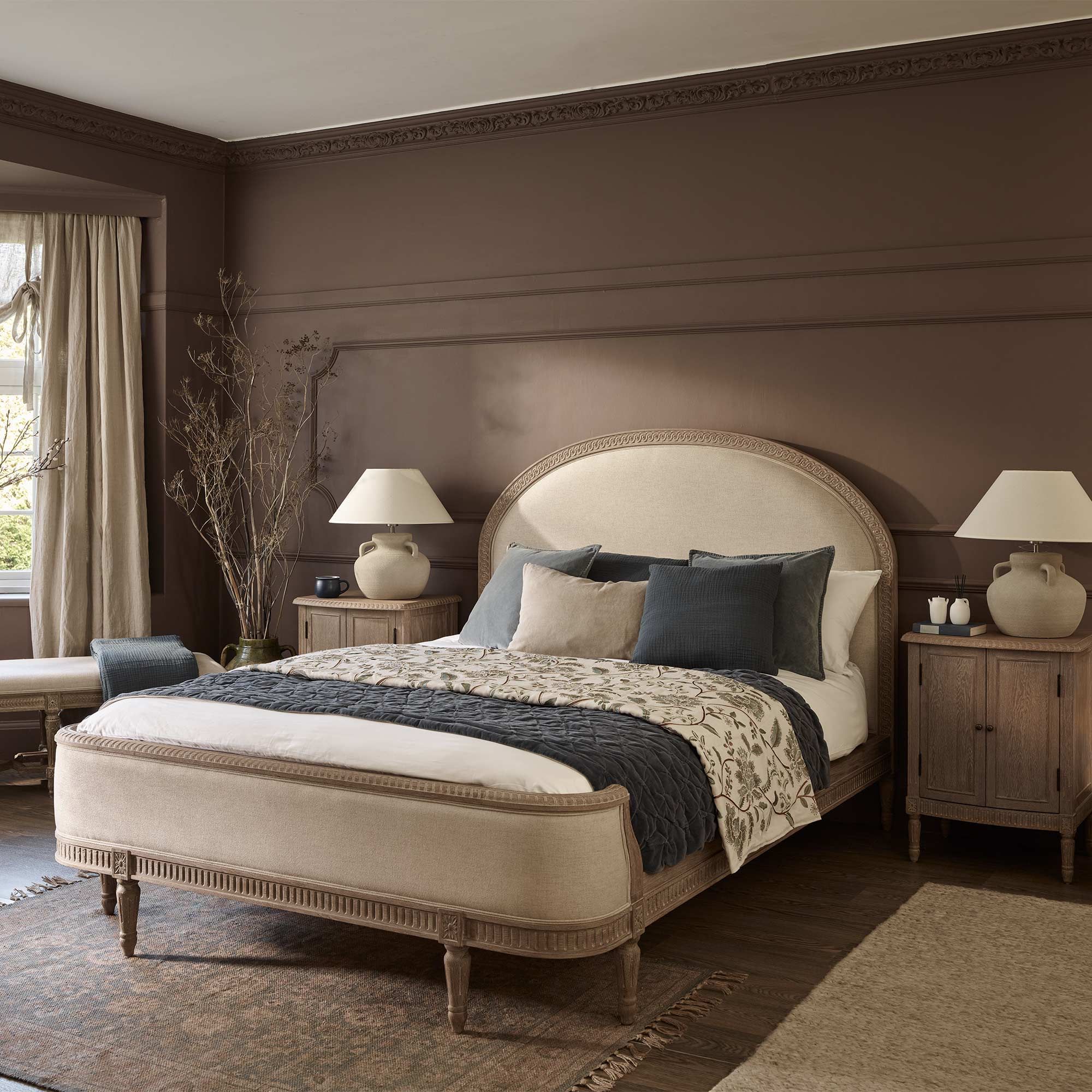 Cozy bedroom with a beige upholstered bed, blue and gray pillows, wooden nightstands, lamps, and a brown accent wall. Large window with curtains.